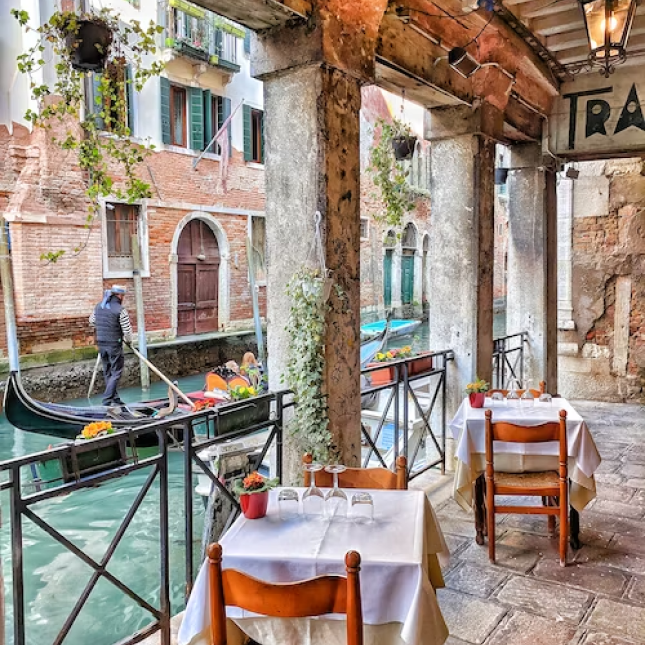 Mesas de restaurante na beira de um canal estreito em Veneza com um gondoleiro passando.